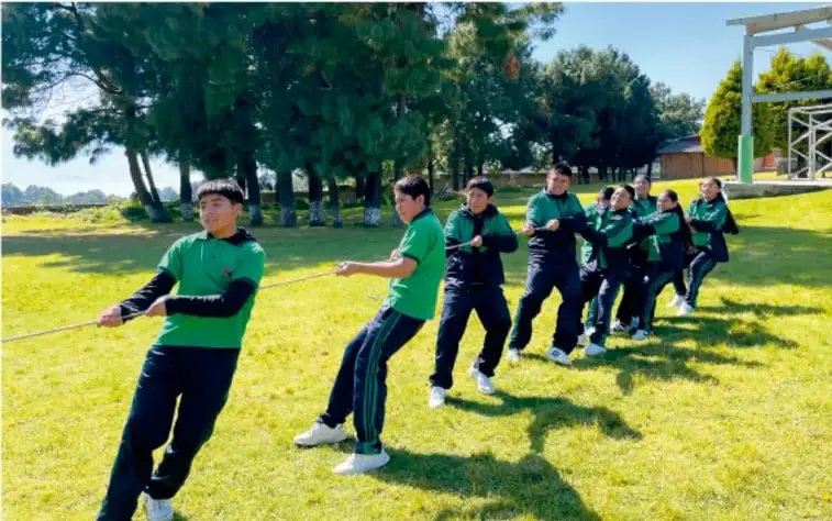 Fotografía de un grupo de estudiantes de secundaria en uniforme verde y azul realizando una actividad de fuerza con una cuerda en un campo de pasto con árboles al fondo, practicando una decisión colectiva sincronizada.