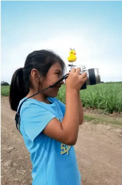 Niña de camiseta azul con un pollito amarillo de juguete sobre la cámara, apuntando con una cámara fotográfica en un campo verde.