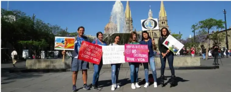 Fotografía de un grupo de jóvenes con pancartas en una plaza pública, frente a una fuente y edificios históricos. Las pancartas aluden a la defensa de los derechos humanos, la no violencia y la construcción de paz.