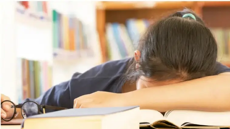 Fotografía de una joven estudiante apoyada sobre su brazo en una mesa de biblioteca, con la cabeza gacha y expresión cansada o triste, rodeada de libros, lo que ilustra la sensación de no encajar en un lugar nuevo.