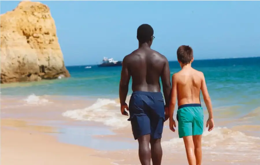 Fotografía de un hombre de piel oscura y un niño de piel clara, de espaldas, caminando juntos por una playa con agua turquesa y rocas al fondo, en actitud de acompañamiento y cariño.