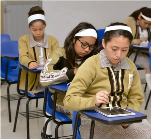 Fotografía de estudiantes de secundaria con uniforme y vineta blanca en la cabeza, sentados en pupitres azules utilizando tablets y dispositivos digitales en el aula.