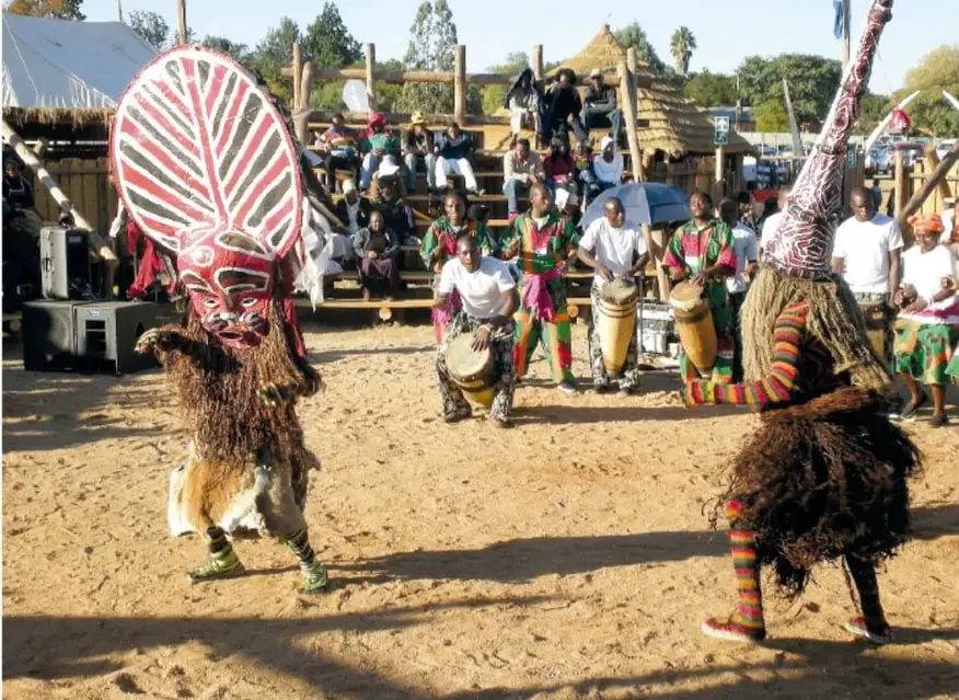 Danzantes con máscaras makishi en un pueblo de Zambia: uno con máscara roja y blanca de rostro humano con penacho de fibra, otro de alto tocado cónico; al fondo, público sentado y músicos con tambores.