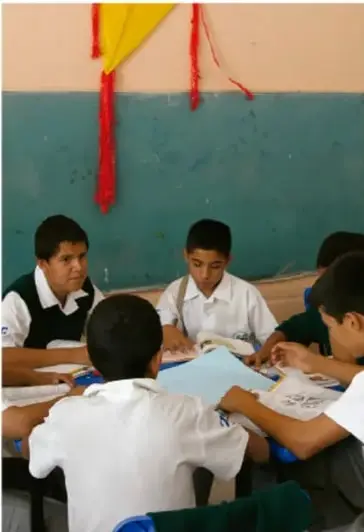 Fotografía de estudiantes de secundaria reunidos en grupo alrededor de una mesa, revisando documentos, en un salón de clases con pizarrón verde.