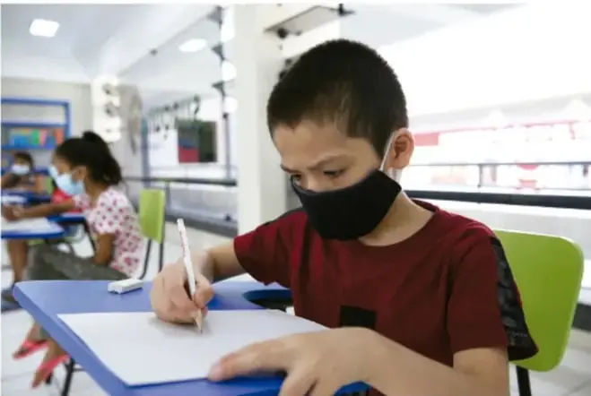 Niño con cubrebocas escribiendo en su cuaderno en un salón de clases; al fondo, una compañera también trabaja.