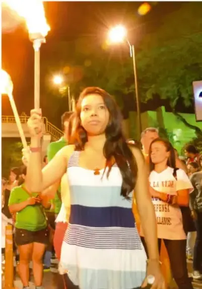 Fotografía de una marcha nocturna ciudadana con antorchas; una joven con camiseta blanca y rayas azules sostiene una antorcha encendida, rodeada de otras participantes. Representa un movimiento social en defensa de derechos.