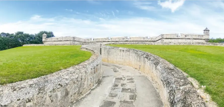 Fotografía de un cañón antiguo sobre cureña de madera dentro de un baluarte con garita abovedada; al fondo se ven palmeras y edificios modernos.