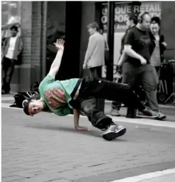 Fotografía de una persona haciendo breakdance o una acrobacia en una calle peatonal con transeúntes al fondo, ilustrando la actividad física.
