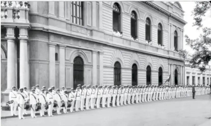 Fotografía en blanco y negro de una formación de marinos uniformados frente a un edificio de arquitectura clásica con columnas y arcos.
