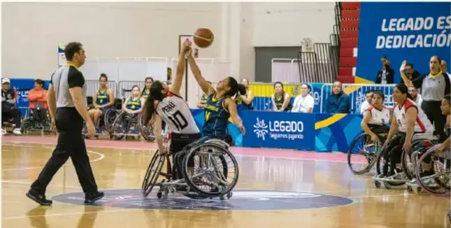 Fotografía de un partido de basquetbol en silla de ruedas entre equipos femeniles, con árbitro y espectadores; al fondo lonas con el texto "Legado es dedicación" (programa Legado).