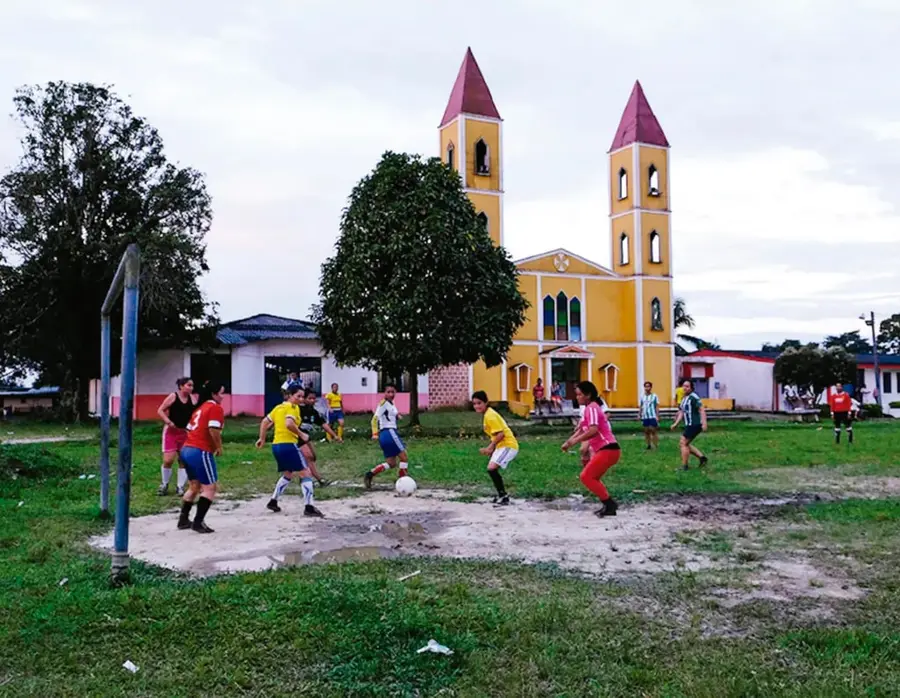 Fotografía de una cancha de fútbol con niños jugando; al fondo se observa una iglesia amarilla con dos torres rojas, árboles, casas y personas alrededor.