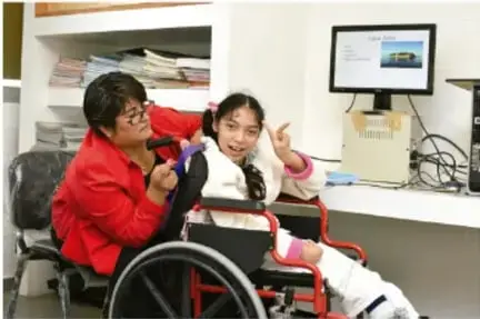 Estudiante de bata blanca presentando una investigación a otro compañero en un aula con computadora.
