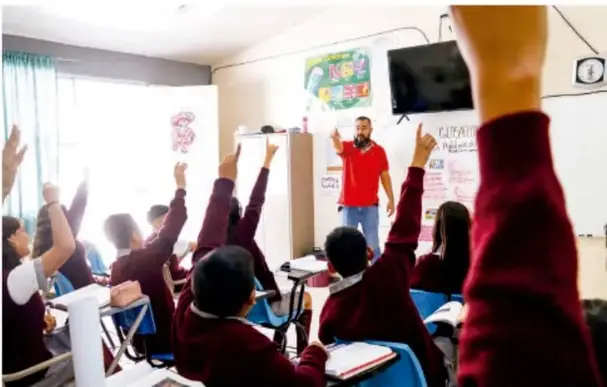 Fotografía de un aula de telesecundaria: un grupo de estudiantes de secundaria con uniforme (camisa blanca y suéter vino) levanta la mano mientras el docente, con camiseta roja, dirige la sesión frente al pizarrón y un televisor.