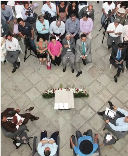 Fotografía aérea de un grupo numeroso de personas sentadas en sillas alrededor de un espacio central con una mesa y arreglos florales, en un patio con piso de piedra. Sugiere una reunión o asamblea comunitaria.