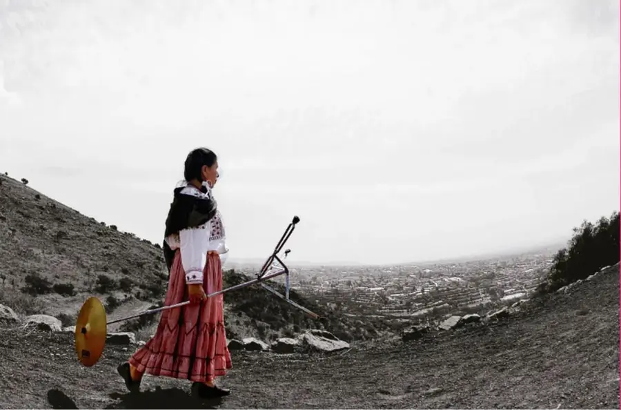 Fotografía en blanco y negro de una mujer indígena cargando un bastón o herramienta en un cerro, con la ciudad al fondo; crédito: Raúl Barajas, Ciudad de México.