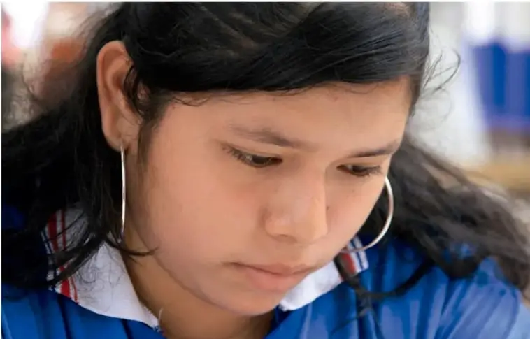 Fotografía de una estudiante de secundaria con cabello largo oscuro, aretes circulares grandes y camisa azul, con la mirada dirigida hacia abajo como si leyera o escribiera.