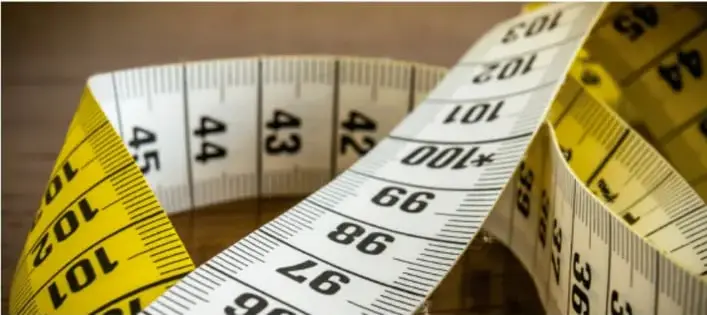 Fotografía de una cinta métrica amarilla y blanca con marcas numéricas visibles (42, 43, 44, 45, 97, 98, 99, 100, 101, 102 cm) enrollada, ilustrando instrumentos de medición de longitud.