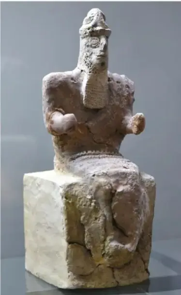 Fotografía de una estatua sumeria de piedra de un dios o gobernante sentado en un trono de cubo, con barba larga y vestimenta, conservada en un museo.