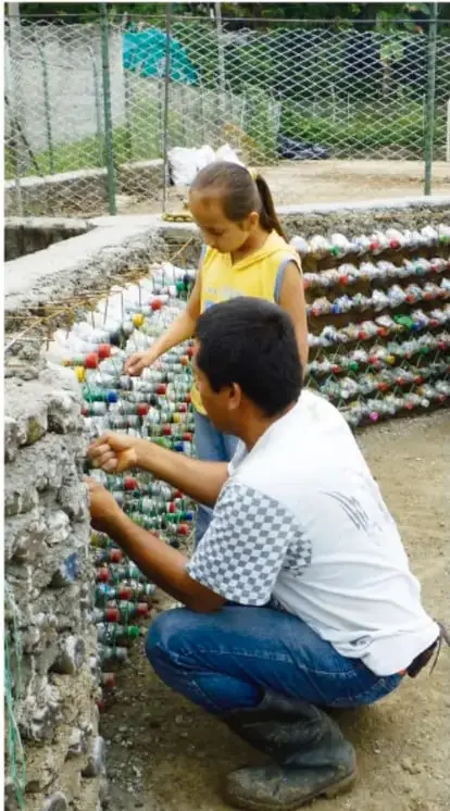 Fotografía de un adulto y una niña colocando ecoladrillos en una construcción con mezcla, dentro de un espacio comunitario con malla ciclónica.