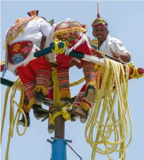 Fotografía de un volador o danzante tradicional con vestimenta colorida de origen indígena, atado con cuerdas a un poste, ilustrando una manifestación cultural tradicional.