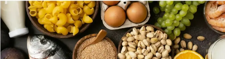 Fotografía de alimentos variados sobre una mesa oscura: botella de leche, pasta, huevos, uvas verdes, camarones, pescado, semillas, granos, naranja y aceite.