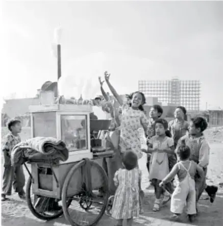 Fotografía en blanco y negro de niños sonrientes alrededor de un carrito callejero, algunos con los brazos alzados.
