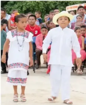 Niña y niño vestidos con trajes tradicionales blancos con bordados de colores y sombrero de palma, participando en una celebración comunitaria entre la gente.