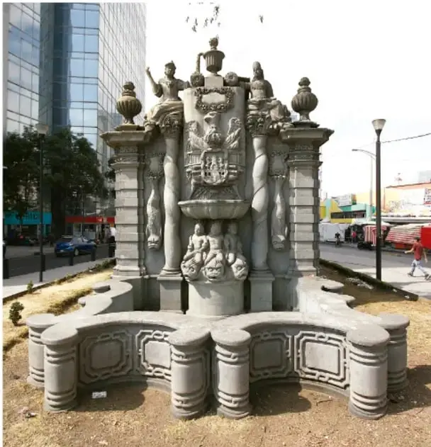 Fotografía de una fuente pública de piedra con esculturas y varios caños, ubicada en una plaza urbana rodeada de edificios, árboles y una calle con vehículos.