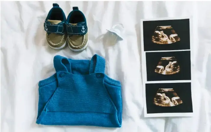 Fotografía de objetos de bebé sobre tela blanca: un par de zapatitos azules, un chupón, un suéter tejido azul y tres ecografías fetales. Representa la planificación y llegada de un bebé.