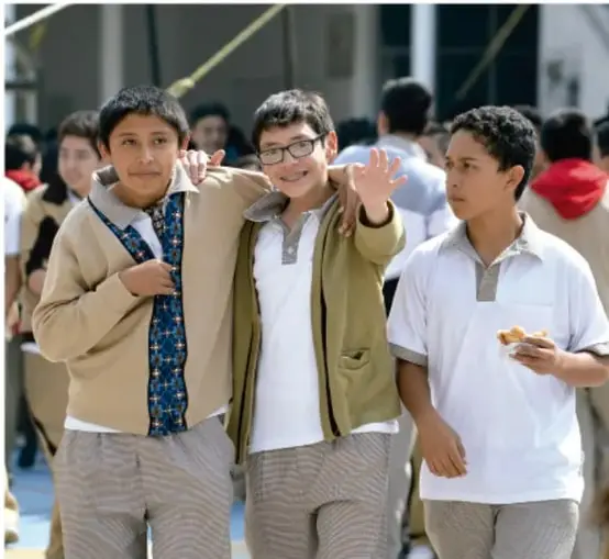 Fotografía de tres adolescentes estudiantes de secundaria en el patio escolar, uno con los brazos abiertos saludando; alrededor hay otros alumnos en actividad.