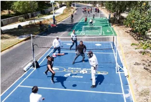 Cancha deportiva pintada de azul en la vía pública con personas jugando voleibol y otros deportes, en un entorno arbolado.