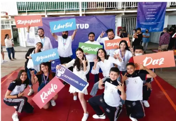 Fotografía de estudiantes y personal de una escuela telesecundaria sosteniendo letreros con nombres de colonias o localidades (Zapopan, Libertad, Arguense, Juventud, Daz, Rabío), posando en el patio escolar durante una actividad comunitaria.