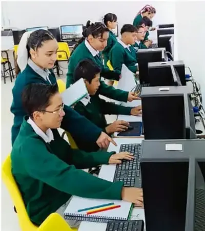 Fotografía de estudiantes de telesecundaria en uniforme verde trabajando en computadoras de la sala de cómputo escolar, organizados en filas.