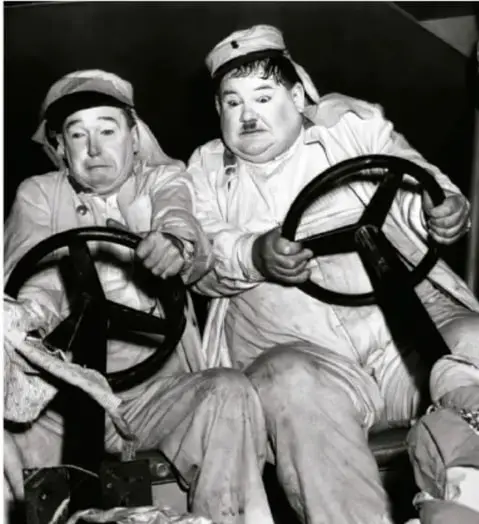 Fotograma en blanco y negro del dúo cómico Laurel y Hardy (El Gordo y el Flaco) sentados ante ruedas de barco o maquinaria, ambos con sombrero.
