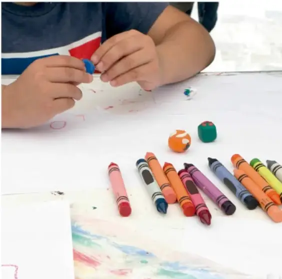 Fotografía de un niño modelando plastilina con las manos, rodeado de crayones de colores sobre una hoja con garabatos de colores.