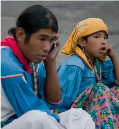 Fotografía de un hombre y una mujer indígenas con vestimenta tradicional; el hombre habla por teléfono celular y la mujer viste blusa azul y pañoleta amarilla.