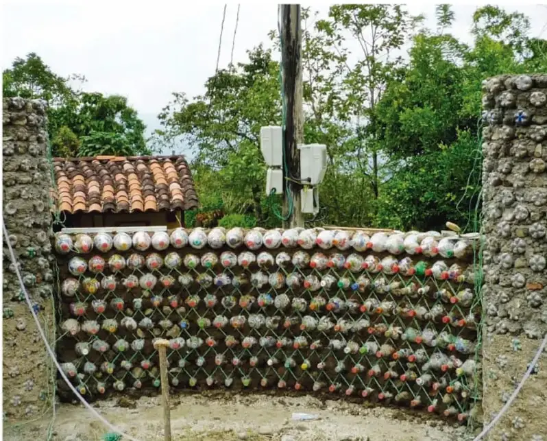 Fotografía de un muro construido con ecoladrillos: botellas de plástico rellenas con residuos de colores, sujetas con una red sobre una base de piedra, frente a un paisaje rural con árboles y una casa de techo de tejas.