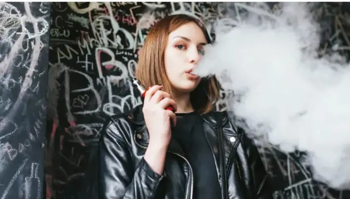 Fotografía de una joven exhala vapor frente a un muro con grafitis, ilustrando el consumo de vapeador.