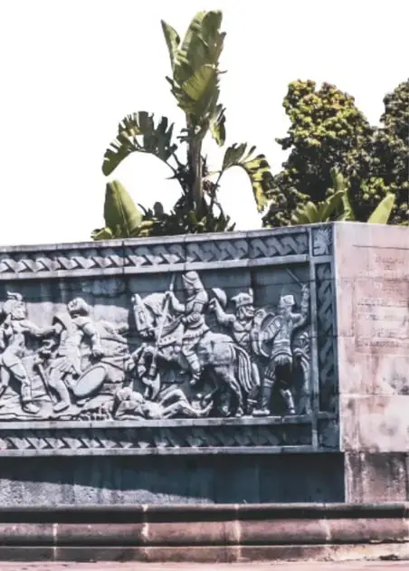 Fotografía de un relieve escultórico del monumento con figuras de conquistadores a caballo, rodeado de vegetación.