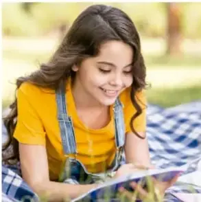 Niña con blusa amarilla y overol de mezclilla sentada en un pasto a cuadros, leyendo un libro.