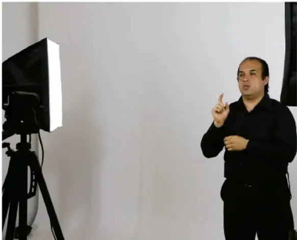 Fotografía de un intérprete de lenguaje de señas, hombre de camisa negra, realizando señas frente a una pantalla y cámara de video sobre trípode, ilustrando la traducción inclusiva.