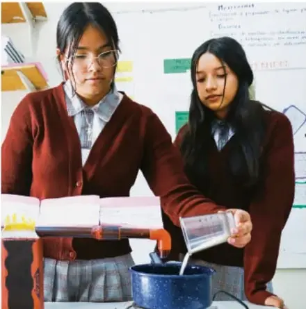 Dos estudiantes en uniforme realizan una práctica de medición vertiendo un líquido desde una probeta en un recipiente sobre una mesa de laboratorio escolar.