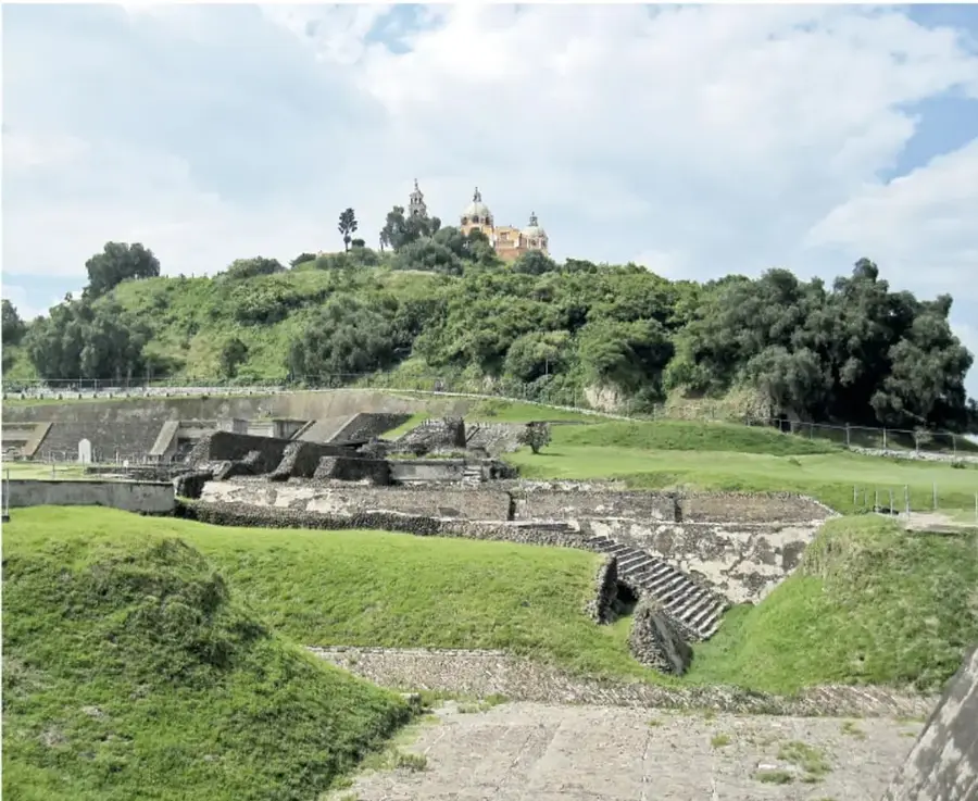 Fotografía panorámica de la Gran Pirámide de Cholula cubierta de vegetación, con la iglesia de Nuestra Señora de los Remedios en la cima y restos arqueológicos de escalinatas y muros en primer plano.