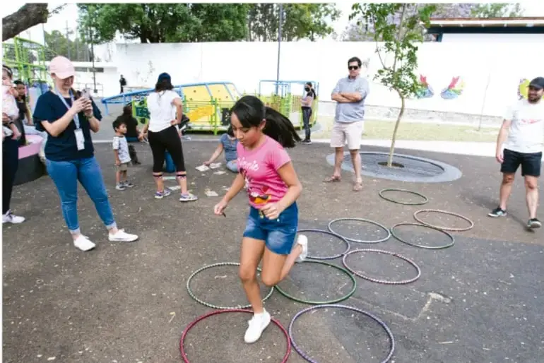 Fotografía de niña saltando entre aros de colores en el patio escolar, con compañeros y adultos observando; actividad física y recreativa al aire libre.