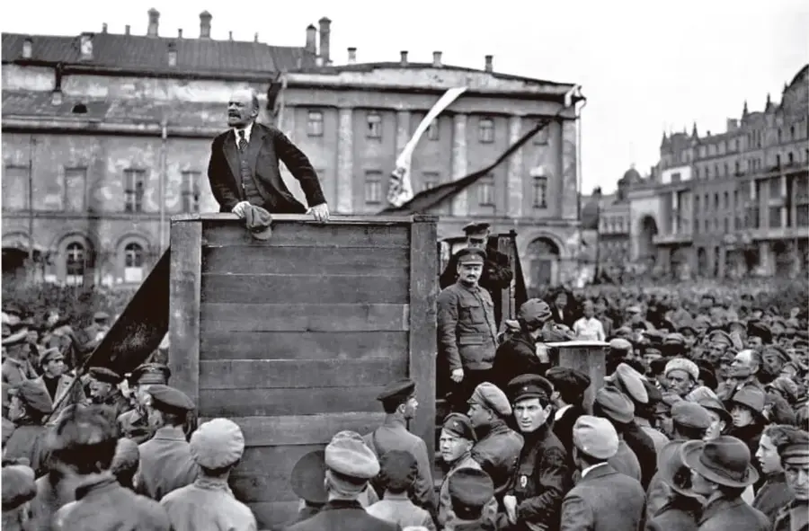 Fotografía en blanco y negro de un líder político (Vladímir Lenin) de pie sobre una tribuna de madera dirigiéndose a una multitud en una plaza pública, con edificios al fondo.