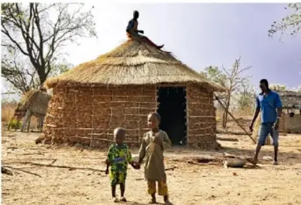 Fotografía de una familia en una comunidad rural con una casa de adobe y techo de paja; un niño y una niña caminan y un adulto observa.