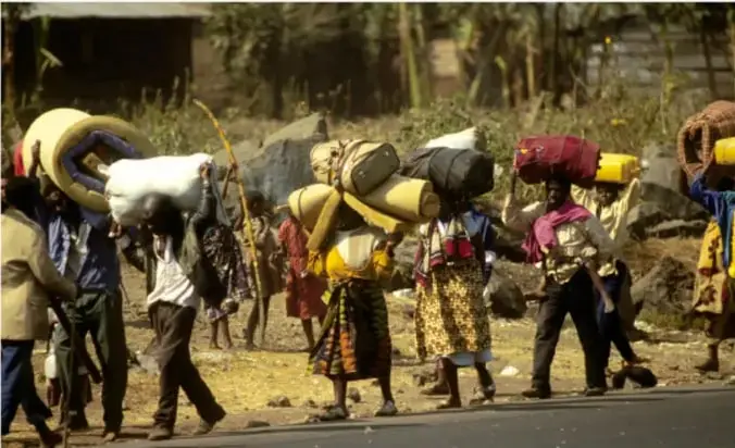Fotografía a color de una columna de personas desplazadas caminando por un camino rural, cargando colchones y bultos sobre la cabeza y la espalda, escena asociada al éxodo durante el genocidio de Ruanda.