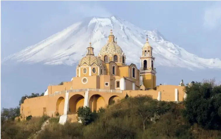 Fotografía de la iglesia de Nuestra Señora de los Remedios, de color amarillo y con cúpulas, construida sobre la cima de la Gran Pirámide de Cholula; al fondo se observa el volcán Popocatépetl cubierto de nieve.