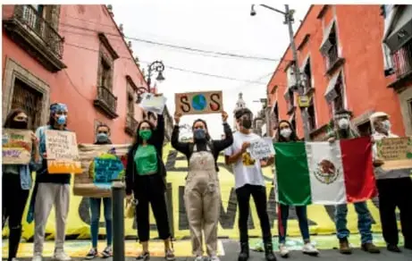 Grupo de personas con cubrebocas sostiene pancartas y banderas en una calle; una pancarta dice "SOS".