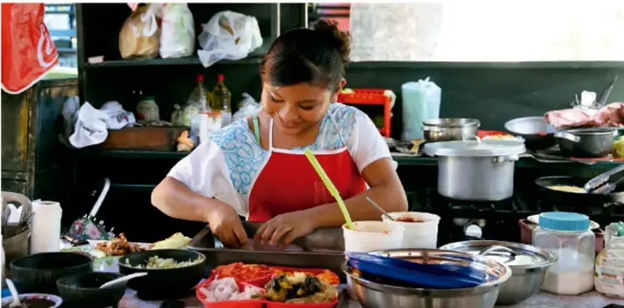 Fotografía de una cocinera con delantal rojo preparando alimentos en una cocina tradicional; sobre la mesa hay ollas, recipientes con ingredientes cortados (cebolla, limón), utensilios y comida en preparación.